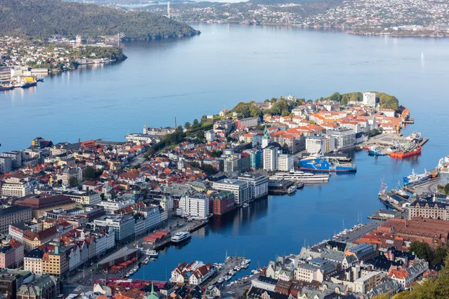 Bergen seen from high above on a clear day, with the Vågen harbour running into the centre, red-roofed streets on the Nordnes peninsula and calm blue water stretching out towards a bridge and wooded hills